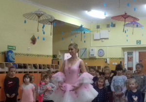 Dziewczynki stoją na środku sali, ćwiczą układ taneczny, prezentowany przez baletnicę.