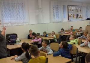 Dzieci siedzą w ławkach. Nauczycielka stoi przed dziećmi.