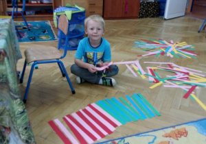 Chłopiec układa na dywanie wzory z pasków z papieru.