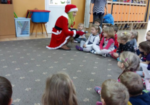 Dzieci siedzą w półkolu podczas zabawy mikołajkowej. Pani Mikołajowa trzyma w reku mikrofon i zadaje dzieciom pytania.