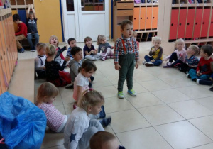 Dzieci siedzą na podłodze w szatni przedszkolnej. W środku stoi chłopiec.