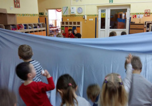 Dzieci biorą udział w konkursie na najszybsze przerzucenie przez niebieski materiał kul z papieru na stronę przeciwnika.