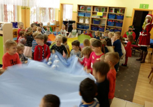 Dzieci w kole trzymając niebieski materiał, podrzucają do góry kulki z papieru