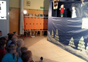 Dzieci siedząc na dywanie oglądają przedstawienie pt. „Opowieść zimowa”. Na scenie widać pacynki w scenerii zimowej