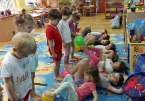 Dzieci podczas zajęć z gimnastyki korekcyjnej wykonują ćwiczenia stóp. Część dzieci leży na dywanie i stopami podaje piłkę stojącym dzieciom piłkę