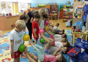 Dzieci podczas zajęć z gimnastyki korekcyjnej wykonują ćwiczenia stóp. Część dzieci leży na dywanie i stopami podaje piłkę stojącym dzieciom piłkę