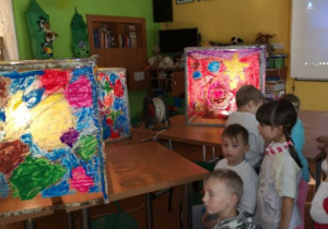 Dzieci oglądają prace plastyczne. Prace przedstawiają sześciany z pomalowanymi w kolorowe wzory ściany. Bryły sa podświetlone.