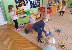 Dzieci bawią się zabawkami na dywanie. W tle widoczne są meble przedszkolne i zabawki.