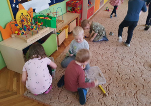 Dzieci bawią się zabawkami na dywanie. W tle widoczne są meble przedszkolne i zabawki.