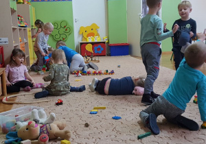 Dzieci bawią się zabawkami na dywanie. W tle widoczne są meble przedszkolne i zabawki.