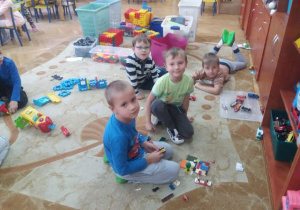 Dzieci siedząc na dywanie, prezentują swoją budowlę z plastikowych klocków