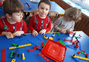 Dzieci siedzą przy stolikach. Budują z plastikowych klocków konstrukcje według własnego pomysłu