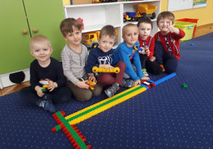 Dzieci siedząc na dywanie, prezentują swoją budowlę z plastikowych klocków