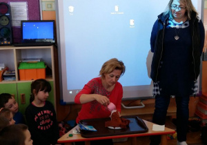 Dzieci siedzą na dywanie. Nauczycielka prezentuje doświadczenie z wulkanem.