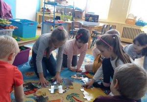 Dzieci układają puzzle na dywanie przedstawiające Układ Słoneczny. W tle widoczne są tablice dydaktyczne, meble przedszkolne i zabawki.