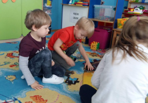Dzieci układają puzzle na dywanie przedstawiające Układ Słoneczny. W tle widoczne są tablice dydaktyczne, meble przedszkolne i zabawki.