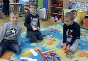 Dzieci układają na dywanie wzory (karmnik) z kolorowych pasków papieru. . W tle widoczne są meble przedszkolne i zabawki.