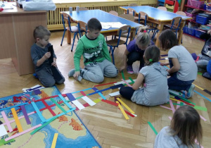 Dzieci układają na dywanie wzory z kolorowych pasków papieru. W tle widoczne są meble przedszkolne i zabawki.