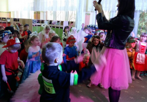 Dzieci przebrane w stroje karnawałowe bawią się na balu. Prowadząca bal trzyma w ręku mikrofon. W tle widać szafki w szatni przedszkolnej.