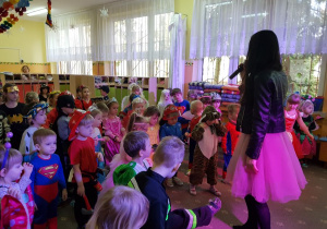 Dzieci przebrane w stroje karnawałowe bawią się na balu. Prowadząca bal trzyma w ręku mikrofon. W tle widać szafki w szatni przedszkolnej.