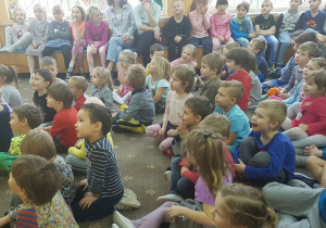 Dzieci siedzą w szatni przedszkolnej na dywanie i oglądają występ muzyka. Zgłaszają się do odpowiedzi.