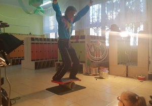 Akrobata stoi w szatni przedszkolnej i prezentuje pokaz ekwilibrystyki: balans na wałku ekwilibrystycznym