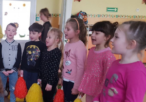 Dzieci stoją w półkolu. W ręku trzymają pompony. Spiewaja piosenkę.