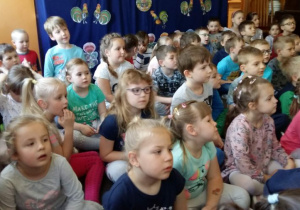 Dzieci siedzą w szatni przedszkolnej na dywanie i oglądają występ aktorów.