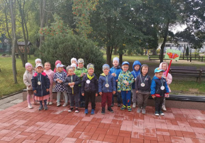 Dzieci 4, 5 - letnie z grupy III stoją na tarasie przedszkolnym w ubraniach jesiennych. Na szyjach mają medale przedszkolaka. Dziewczynka trzyma symbol motyla na tyczce.