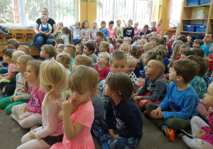 Dzieci siedzą w szatni przedszkolnej na dywanie i oglądają występ muzyka.