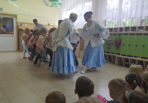 Przedszkolaki stoją na środku sali, ćwiczą układ taneczny, prezentowany przez artystki. W tle widać szafki w szatni przedszkolnej.