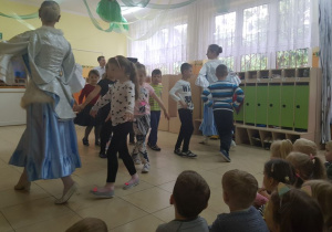 Przedszkolaki stoją na środku sali, ćwiczą układ taneczny, prezentowany przez artystki. W tle widać szafki w szatni przedszkolnej.