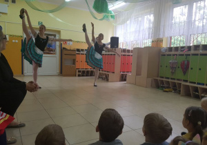 Dzieci siedzą na dywanie. Przed nimi na scenie w szatni przedszkolnej tańczą 2 baletnice. W tle widać szafki w szatni przedszkolnej.