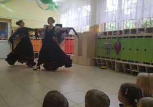 Dzieci siedzą na dywanie. Przed nimi na scenie w szatni przedszkolnej tańczą 2 baletnice, ubrane w czarne suknie. W tle widać szafki w szatni przedszkolnej.