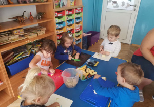 Dzieci 4, 5 - letnie siedzą przy niebieskim stole, kroją banany jabłka i gruszki plastikowymi nożami.