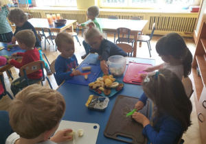 Dzieci 4 - letnie siedzą przy niebieskim stole, kroją banany, jabłka i gruszki plastikowymi nożami. Dziewczynka sięga po pomarańczę.