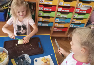 Dzieci 4 - letnie siedzą przy niebieskim stole i kroją brzoskwinie i gruszki plastikowymi nożami