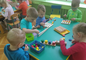 Dzieci siedzą przy stolikach. Uczestniczą w zabawach sensorycznych. Na stolikach rozłożone są pomoce Montessori.