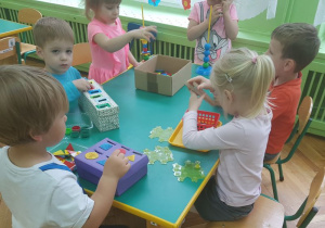 Dzieci siedzą przy stolikach. Uczestniczą w zabawach sensorycznych. Na stolikach rozłożone są pomoce Montessori.