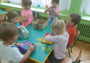 Dzieci siedzą przy stolikach. Uczestniczą w zabawach sensorycznych. Na stolikach rozłożone są pomoce Montessori.