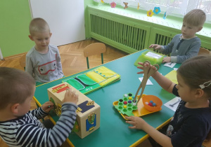 Dzieci siedzą przy stolikach. Uczestniczą w zabawach sensorycznych. Na stolikach rozłożone są pomoce Montessori.