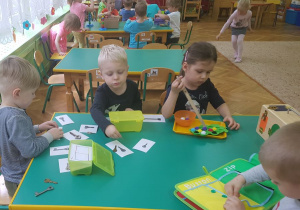 Dzieci siedzą przy stolikach. Uczestniczą w zabawach sensorycznych. Na stolikach rozłożone są pomoce Montessori.