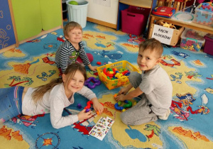 Dzieci na dywanie bawią się klockami. W tle widoczne są meble przedszkolne, tablice dydaktyczne i zabawki.