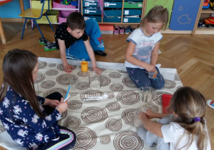Dzieci siedzą na podłodze, blisko rozłożonej ceraty, na której przesypują ryż za pomocą plastikowych miarek. W tle widoczne są meble przedszkolne, tablice dydaktyczne i zabawki.