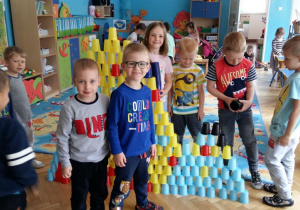 Dzieci układają konstrukcje z plastikowych, kolorowych kubków. W tle widoczne są meble przedszkolne.