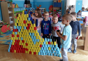 Dzieci układają konstrukcje z plastikowych, kolorowych kubków. W tle widoczne są meble przedszkolne.
