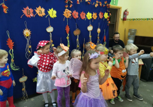 Dzieci przebrane w jesienne liście i postacie tańczą na granatowym tle z jesienną dekoracją kaczuszki.