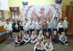 Dzieci z grupy III i V na tle biało - czerwonej dekoracji. Dzieci mają przypiete własnoręcznie wykonane kotyliony.