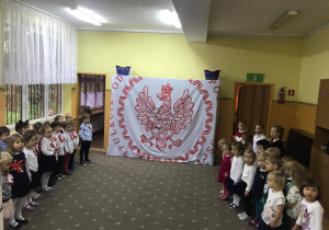 Dzieci stoją na baczność i śpiewają hymn narodowy. W tle biało-czerwona dekoracja z Orłem Białym