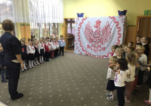Dzieci stoją na baczność i śpiewają hymn narodowy. W tle biało-czerwona dekoracja z Orłem Białym. Nauczycielka prezentuje postawę, którą przyjmuje sie podczas śpiewania hymnu.